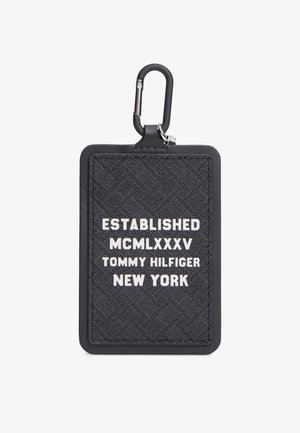 Must tekstuuriga ristkülikukujuline võtmehoidja valge tekstiga, kus on kiri "Established MCMLXXXV Tommy Hilfiger New York", ja must karabiinkinnitus.