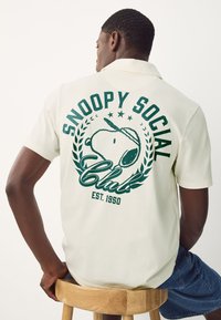 Cremefarbendes Poloshirt mit kurzen Ärmeln, das ein grünes Snoopy-Motiv, großen Text, ein Lorbeer-Design und das Gründungsjahr 1950 auf der Rückseite zeigt.