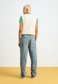 Gilet crema a maglia con motivo a treccia sopra una maglietta verde, abbinato a jeans larghi blu chiaro con molte tasche e un leggero sbiadimento.