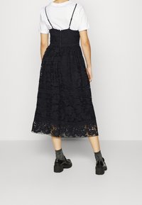 Robe en dentelle noire avec des bretelles spaghetti réglables, motifs floraux et une jupe fluide mi-longue. Portée par-dessus un t-shirt blanc et associée à des chaussures noires imposantes.