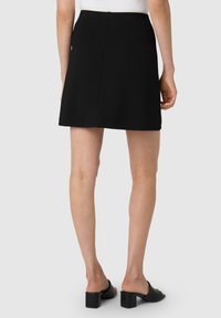Mini gonna nera con una superficie liscia, mostrata da dietro. Presenta una zip nascosta e una vestibilità dritta. Abbinata a scarpe con tacco blocco nere.
