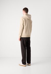 Felpa con cappuccio beige dal tessuto liscio, dotata di tasca a marsupio e polsini a costine, abbinata a pantaloni neri e sneakers bianche.