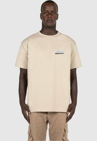 Beige bomull t-skjorte med korte ermer, med en liten grafisk logo i svart og blått på venstre brystområde. Enkle design, avslappet passform.
