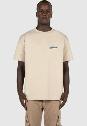 Beige bomull t-skjorte med korte ermer, med en liten grafisk logo i svart og blått på venstre brystområde. Enkle design, avslappet passform.