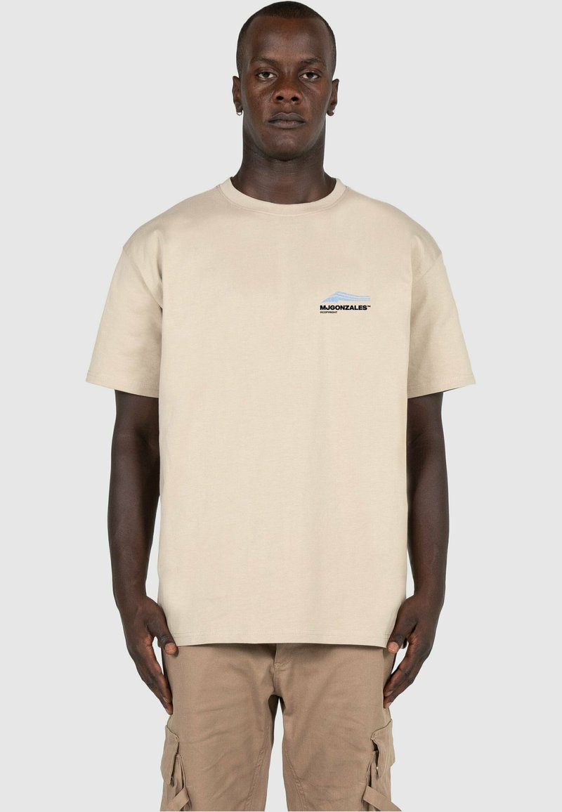Beige bomull t-skjorte med korte ermer, med en liten grafisk logo i svart og blått på venstre brystområde. Enkle design, avslappet passform.