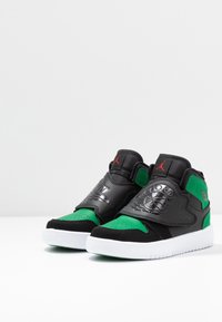 Des baskets montantes noires et vertes avec une tige en cuir et en daim, présentant une sangle latérale à motifs et une semelle en caoutchouc blanche.