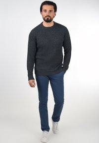 Mørkeblå strikket genser med et teksturert mønster, kombinert med smale blå bukser og hvite sneakers. Genseren har rund hals og ribbestrikkede ermer.