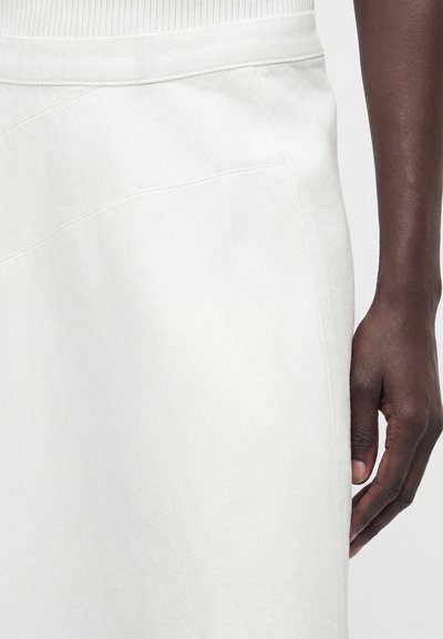 Σκούρο δερμα εκει χέρι που ακουμπά δίπλα σε ένα λευκό φούστα με ψηλή μέση, διαγώνια ραφή και λεπτή ριμπ υφή.