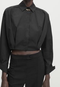 Schwarzes, verkürztes Shirt mit spitzem Kragen, das eine metallische Kettenakzentuierung am Ausschnitt aufweist. Glatte Textur, lange Ärmel mit Manschetten.