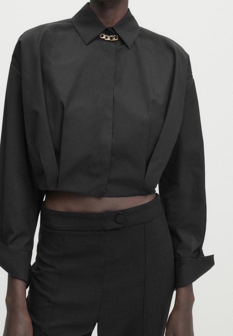 Schwarzes, verkürztes Shirt mit spitzem Kragen, das eine metallische Kettenakzentuierung am Ausschnitt aufweist. Glatte Textur, lange Ärmel mit Manschetten.