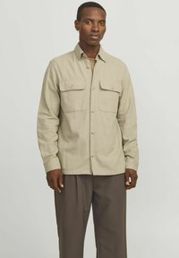 Camicia beige chiaro a maniche lunghe con due tasche sul petto, chiusura a bottoni e tessuto testurizzato. Indossata sopra una camicia chiara, abbinata a pantaloni scuri.