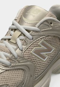 Beiger Laufschuh mit Obermaterial aus Mesh und Synthetik, weißen Schnürsenkeln und strukturierter Oberfläche. Mit einem dezenten Akzent auf dem "N"-Logo.