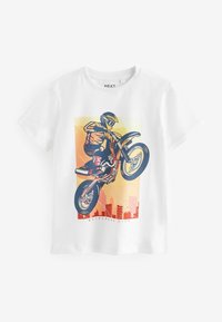 Balts kokvilnas T-krekls ar krāsainu motocross grafiku, kurā braucējs izpilda triku, fons ir pilsētas ainava ar gradientu.
