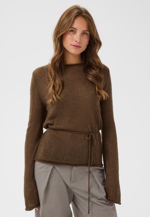 Jeune femme aux longs cheveux ondulés porte un pull marron à manches longues avec une ceinture à la taille et un pantalon gris à carreaux, debout devant un fond uni.