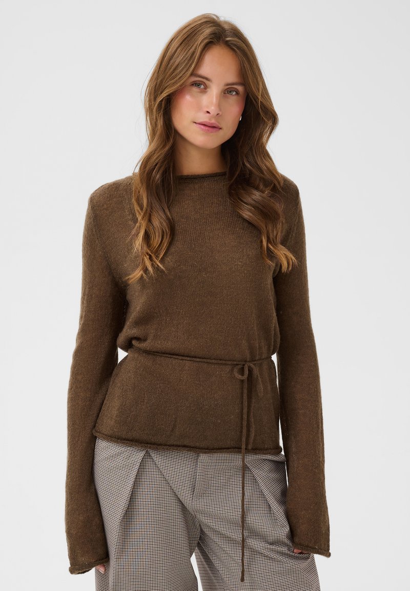Jeune femme aux longs cheveux ondulés porte un pull marron à manches longues avec une ceinture à la taille et un pantalon gris à carreaux, debout devant un fond uni.