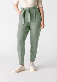 Pantaloni a sigaretta verde chiaro realizzati in un tessuto liscio, con cintura a fiocco, pieghe e tasche laterali, abbinati a sneakers bianche.