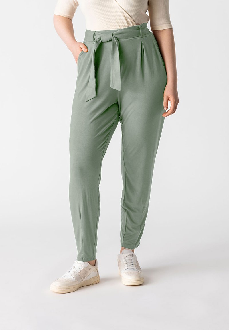 Pantaloni a sigaretta verde chiaro realizzati in un tessuto liscio, con cintura a fiocco, pieghe e tasche laterali, abbinati a sneakers bianche.