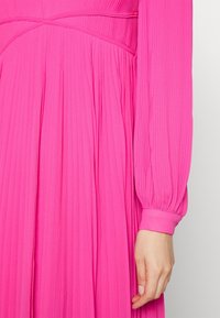 Robe plissée rose avec manches longues à poignets froncés et corsage ajusté. Tissu lisse avec plis verticaux sur toute la surface.