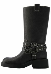Bota hasta la rodilla de cuero sintético negro con tacón grueso, que presenta dos correas decorativas con hebillas y detalles de ojales metálicos. Textura lisa.