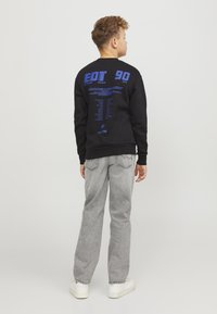 Felpa nera con testo blu sulla schiena, con le parole "EDT 90". Abbinata a jeans grigi chiari e sneakers bianche.