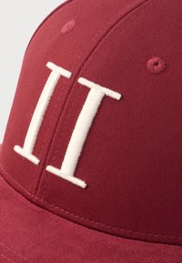 Sarkana kokvilnas beisbola cepure. Priekšpusē ir balta izšūta "II" zīme, ar gludu, teksturētu virsmu un stingru malu.