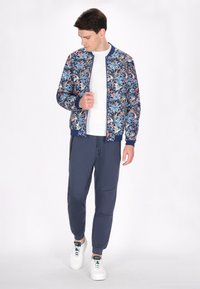 Blommig bomberjacka med blå, vita och rosa mönster; åtsittande, stickade muddar och midja. Kombinerad med marinblå joggers och vita sneakers.