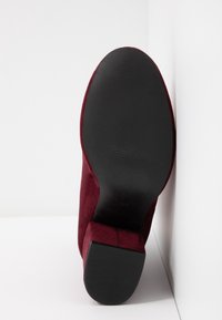 Vue de la semelle d'une botte cheville en daim bordeaux avec un talon bloc, placée à la verticale contre un coin de mur blanc.