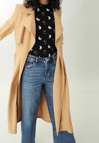 Beige dlouhý trenčkot přes černý top s bílým vzorem jin a jang, spárovaný s jeansy s vysokým pasem v modré barvě. Měkká textura, volný střih.
