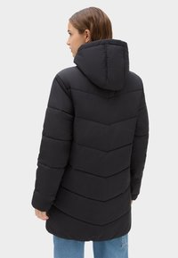 Abrigo acolchado negro con capucha, que presenta un patrón de acolchado en forma de chevron, textura suave y un corte más largo, combinado con jeans de color azul claro.