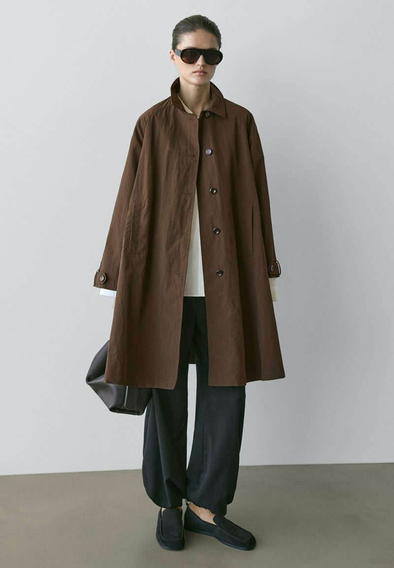 Woman wearing oversized brown buttoned coat, black wide-leg pants, black loafers, sunglasses, and holding a dark handbag, standing on gray floor.