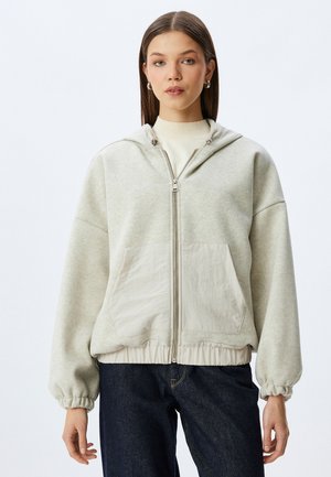 Jasnoszary, rozpinany hoodie o miękkiej fakturze, w oversize'owym kroju i elastycznych mankietach; posiada dużą przednią kieszeń oraz regulowany kaptur.