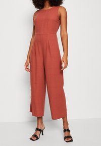Jumpsuit w kolorze rdzy z lnu, bez rękawów, w szerokim kroju, z plisami z przodu i gładką teksturą. Podkreślony czarnymi sandałami na obcasie.