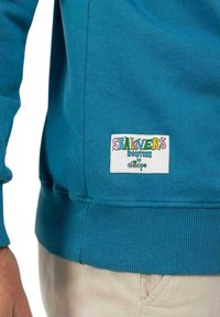 Sudadera verde azulado con puños y dobladillo acanalados, que presenta un parche de marca colorido que dice "SEALOVERS ROUTINE x el tipo."