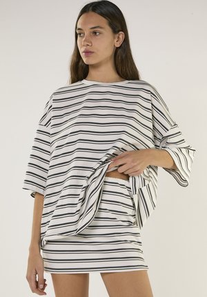 Junge Frau trägt ein weiß-schwarz horizontal gestreiftes, übergroßes Hemd und passenden Rock, hebt das Hemd leicht an, um die Taillendetails zu zeigen.