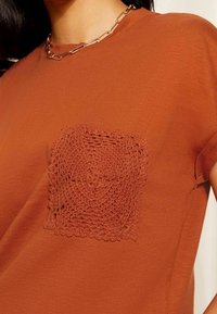 Tričko s rustikálně oranžovou barvou a kulatou háčkovanou nášivkou na levé straně hrudi. Měkká látka, krátké rukávy a uvolněný střih. Viditelný řetízkový náhrdelník.