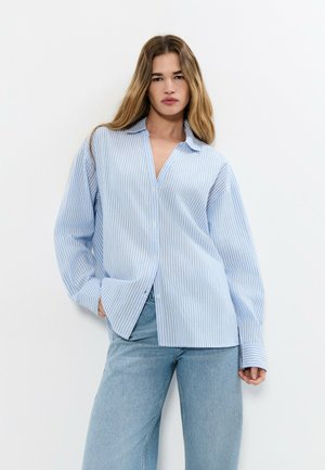 Mujer joven con cabello largo, vistiendo una camisa suelta de rayas azul claro y blanco abotonada y pantalones vaqueros anchos azul claro, sobre un fondo blanco.