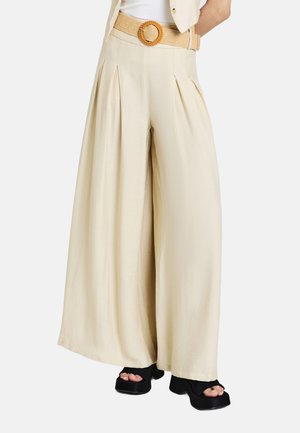 Beige weite Hosen mit Falten, tailliertem Bund und einem strukturierten, geflochtenen Gürtel mit runder Schnalle. Schwarze Plateausandalen runden das Outfit ab.