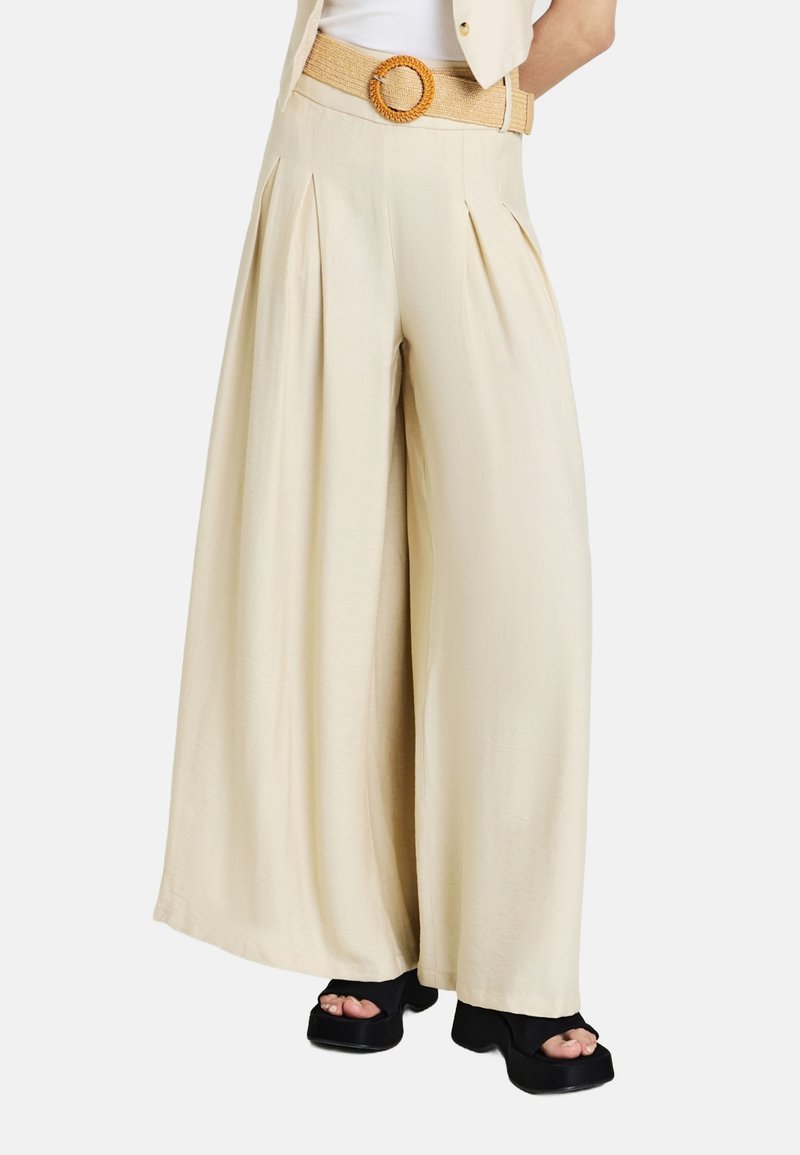 Pantaloni beige a gamba larga con pieghe, vita aderente, e una cintura testurizzata intrecciata con fibbia rotonda. Sandali neri con zeppa completano il look.