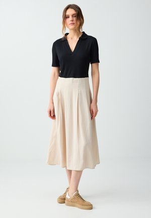 Vrouw staat, draagt een zwart kortemouwen top, een beige midirok en bruine sneakers tegen een lichte rustige achtergrond.