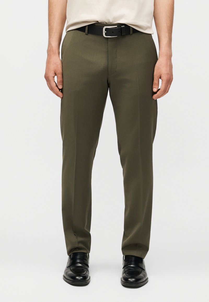 Άνδρας που φοράει ελαιδί παντελόνι, μαύρη δερμάτινη ζώνη και μαύρα επίσημα παπούτσια, στέκεται μπροστά σε απλό φωτεινό φόντο.