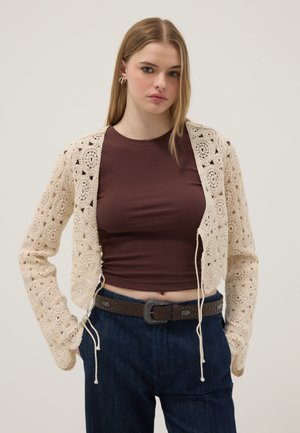 Jonge vrouw die een gebroken witte gehaakte cardigan draagt over een bruin cropped topje en donkerblauwe spijkerbroek met een bruine riem, staand met handen in de zakken.