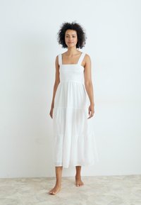 Balta medvilnės maksi suknelė su kvadrato formos iškirpte, surinktu liemeniu ir sluoksniuota sijonine dalimi. Turi plačias petnešas ir laisvą, plazdančią formą.