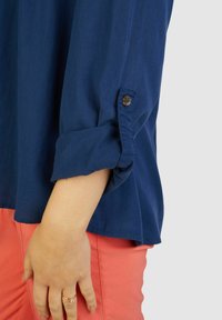 Marineblaue Bluse mit hochgerollten Ärmeln und einem Knopfdetail. Kombiniert mit koralfarbenen Hosen, die eine glatte Stofftextur zeigen.