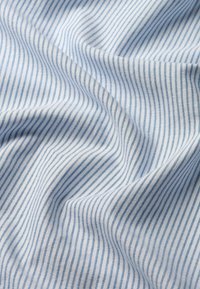 Vaaleansininen ja valkoinen puuvillakangas, jossa on vinoraitoja, erottuu hienolla tekstuurilla ja hienovaraisella kiillotuksella, laskeutuu pehmein poimuin.