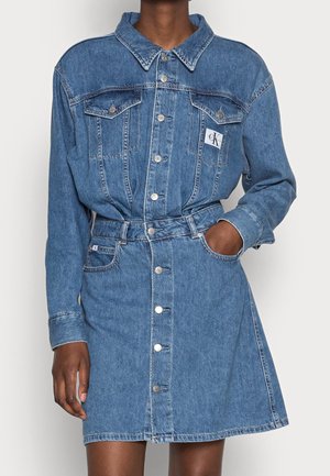 Persona con vestido vaquero azul abotonado, mangas largas, bolsillos en el pecho y una etiqueta de marca en el lado izquierdo, mano en el bolsillo derecho.