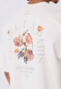 Camiseta blanca con bordado floral colorido que incluye el texto "PROJECT X PARIS" y "WILD SPIRIT PXP COLLECTION" en gris y negro.