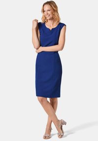 Blaues, tailliertes Kleid mit kurzen Ärmeln, V-Ausschnitt und klaren Linien. Kombiniert mit hellgrauen, offenen Sandalen mit Absatz, die einen runden Akzent an den Riemen haben.