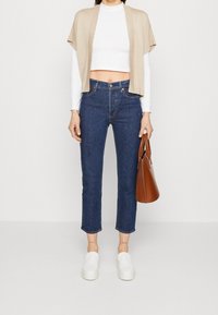 Cardigan corto beige sopra una maglietta a maniche lunghe bianca, abbinato a jeans attillati blu scuro e scarpe slip-on bianche, mentre tiene una borsa marrone.