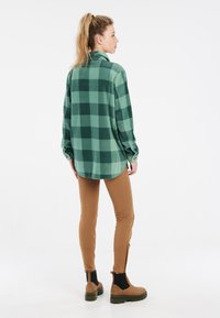 Groen en donkergroen geruit flanellen shirt met knoopmanchetten, gecombineerd met bruine slim-fit broek en bruine enkelboots met zwarte accenten.