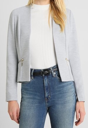 Femme portant un blazer gris clair zippé sur un haut blanc côtelé, associé à un jean bleu taille haute et une ceinture noire.
